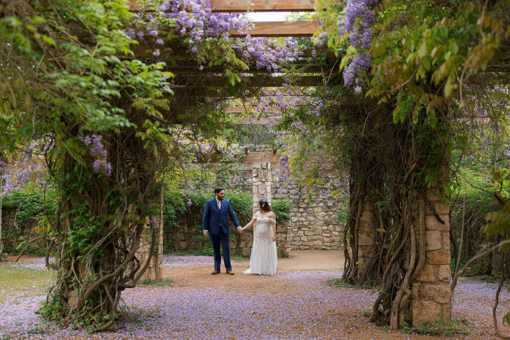 Couple standing under wooden gazebo with wisteria covered pillars surrounded by purple wisteria petals