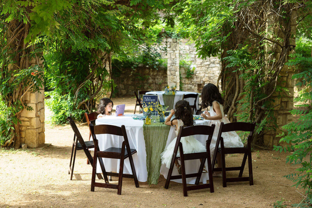 Flowers girls sitting at round table under a wood and stone gazebo covered in vines