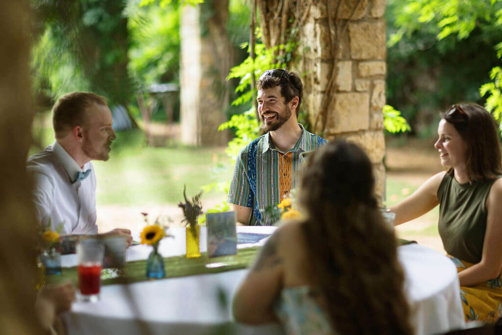 Candid photo of guests laughing while sitting at round table under a wood and stone gazebo covered in vines