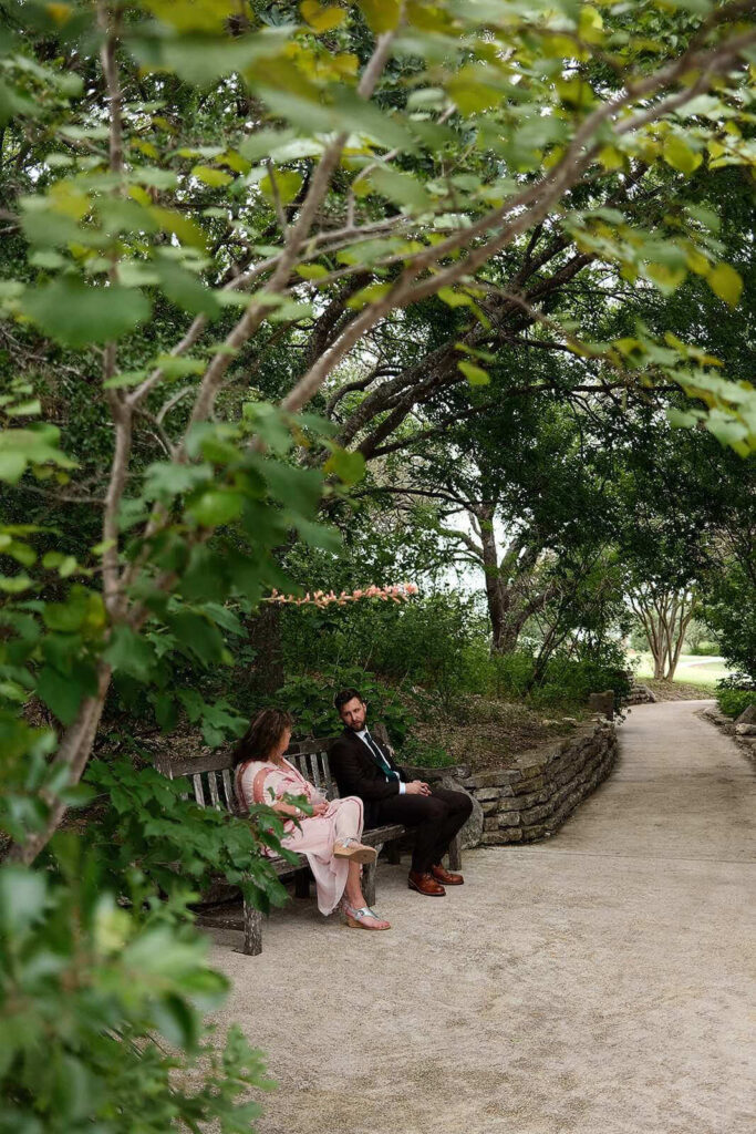 Candid photo of groom sitting on bench with mom framed by trees