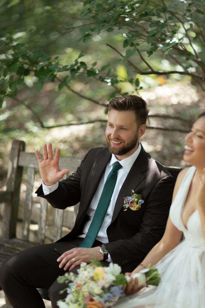 Candid photo of groom sitting on bench and waving at someone while bride laughs in the background