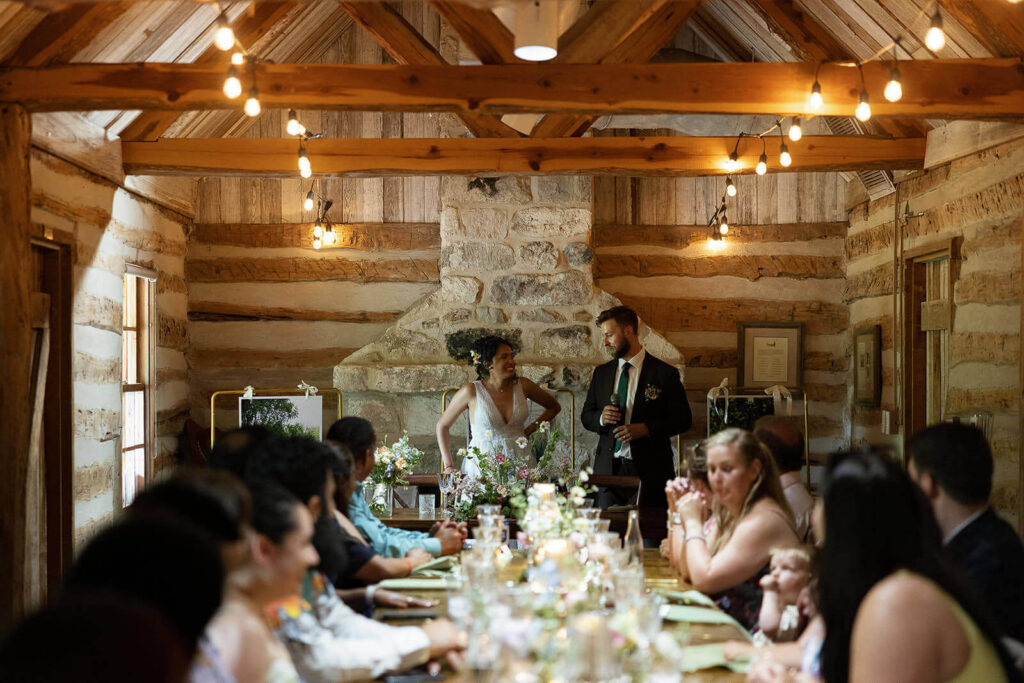 Candid of couple standing at the head of a dinner table lit by string lights and candles inside Auld House at The San Antonio Botanical Garden