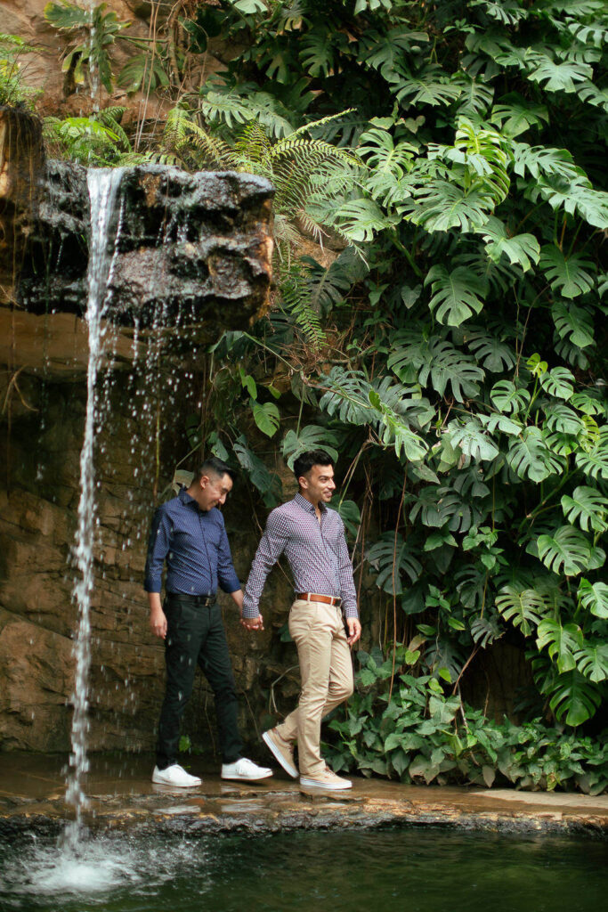 A same sex couple walking under a waterfall surrounded by monstera