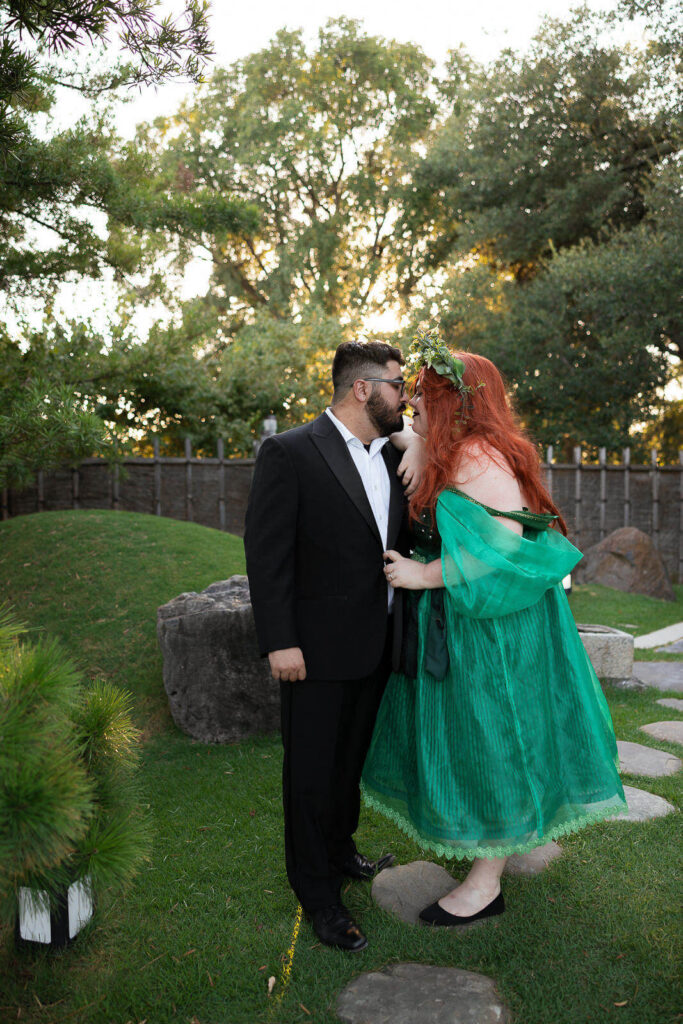 Couple in black suit and green dress rubbing their noses together and standing on a stone path surrounded by grass