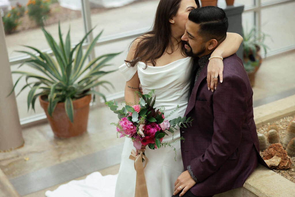Bride sitting in grooms lap whispering in his ear and holding pink and green bouquet at the San Antonio Botanical Garden