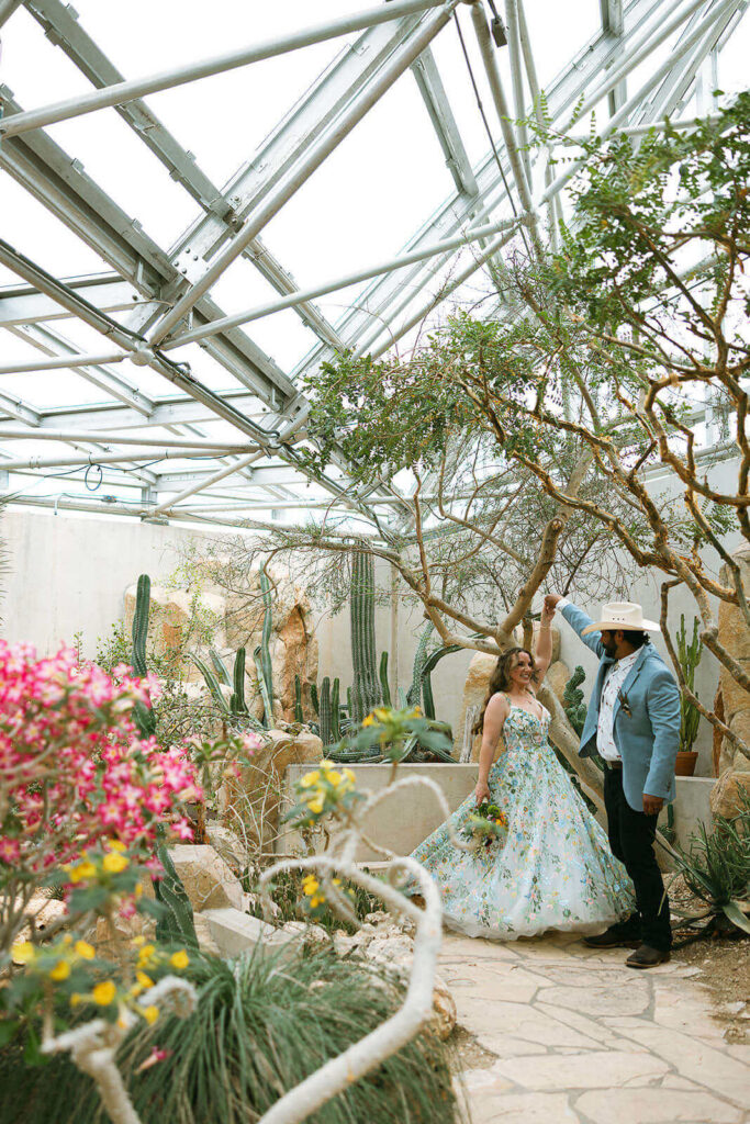Groom in cowboy hat spinning bride in blue embroidered wedding dress surrounded by cacti and desert rose blooms at San Antonio Botanical Garden
