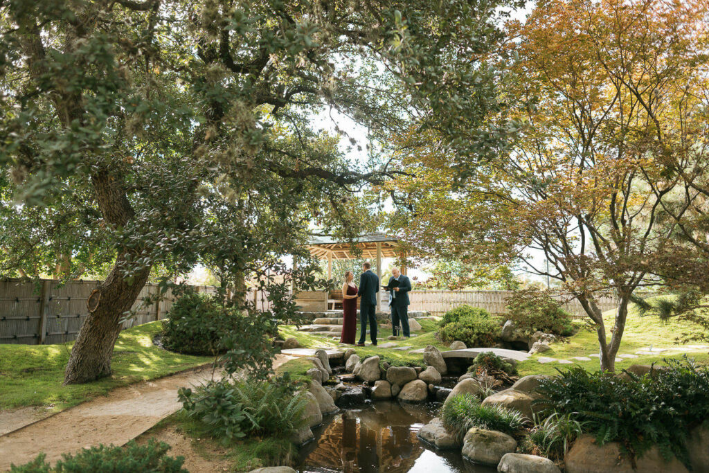 Couple and their officiant exchanging vows in the center of Kumamoto En surrounded by trees, a gazebo, and a pond filled with koi fish