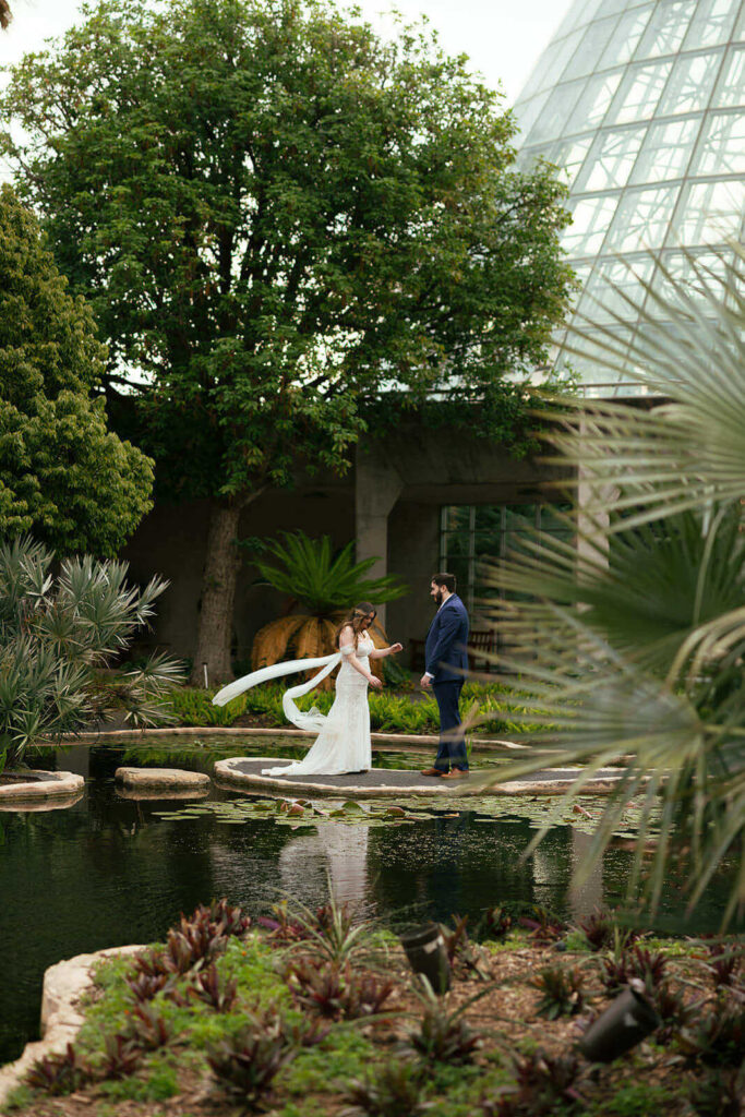 Candid of a bride and groom standing on a concrete island in the middle of a pond while the wind picks up the brides veil