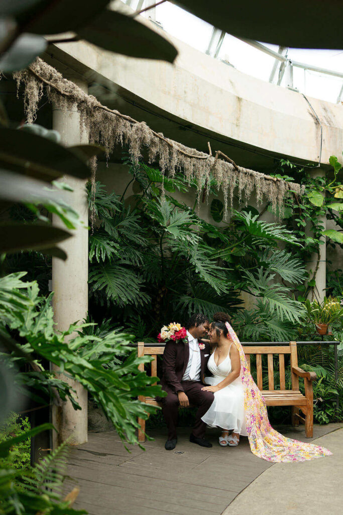 Bride and groom with colorful sequin veil sitting on a bench inside the orchid pavilion
