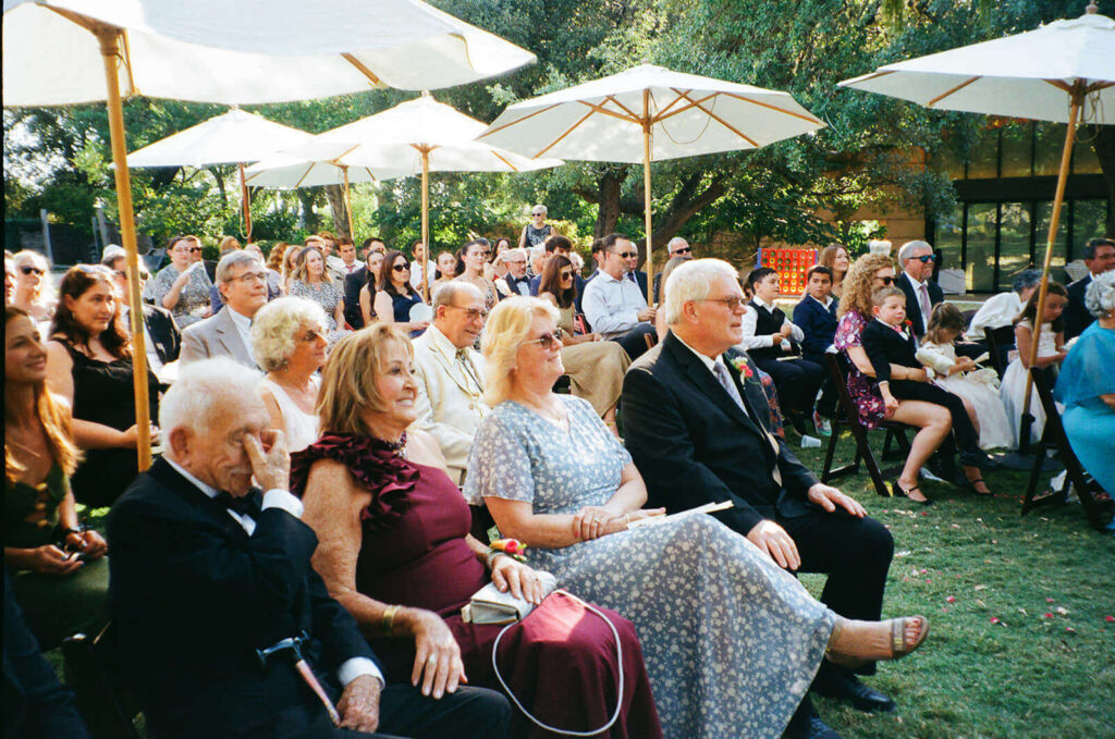 Older guests seated at a wedding ceremony smiling surrounded by umbrellas on 35mm film
